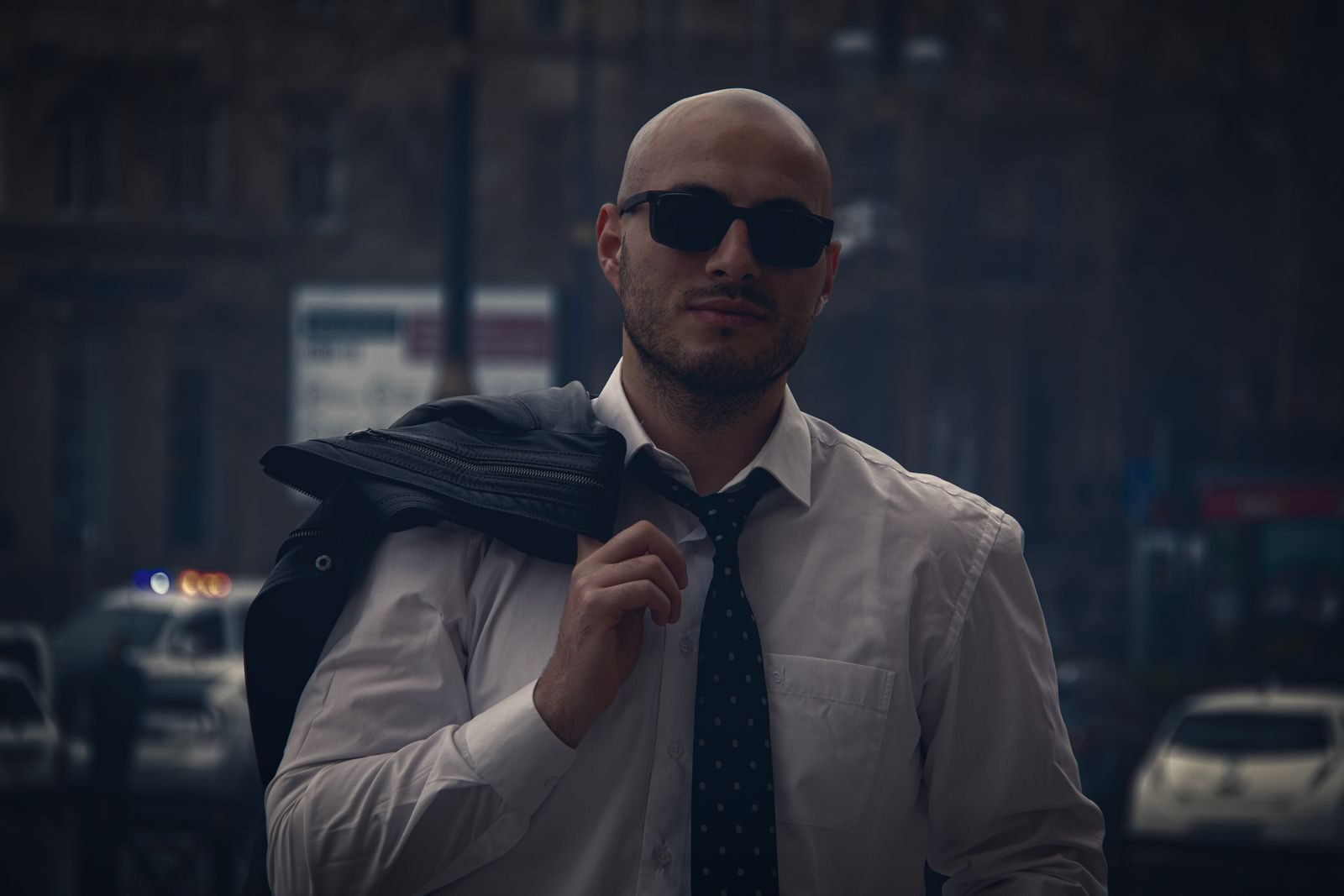 A confident bald man in sunglasses and necktie stands outdoors, exuding urban style.
